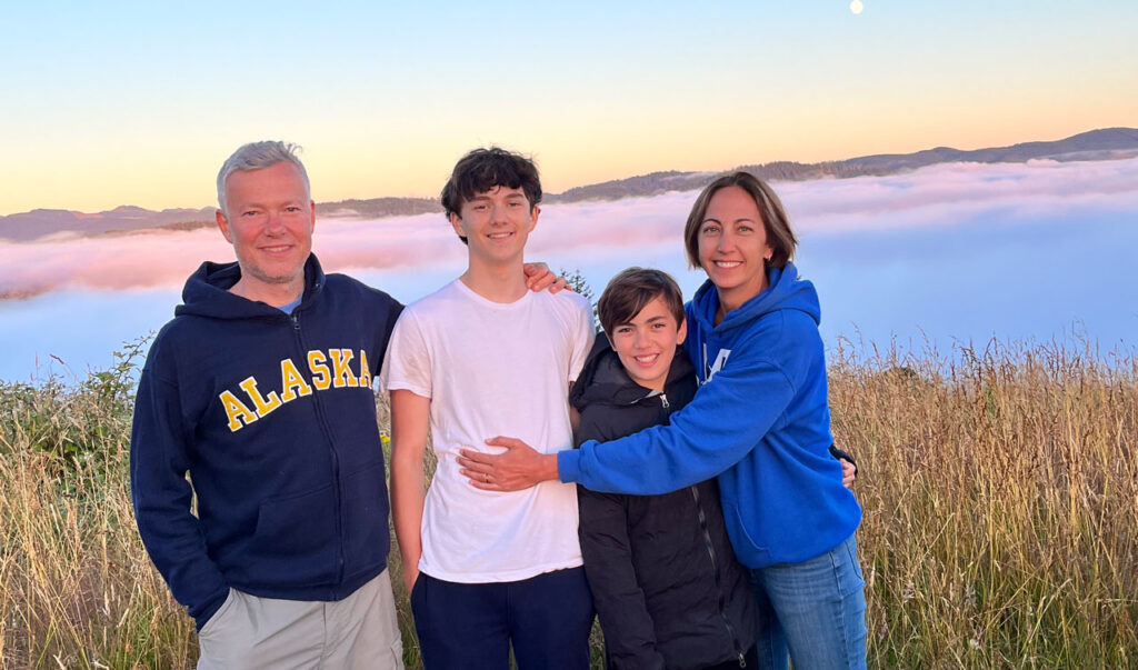 A family of four standing together in a picturesque outdoor setting with a backdrop of clouds and rolling hills. The image captures a warm and joyful moment.